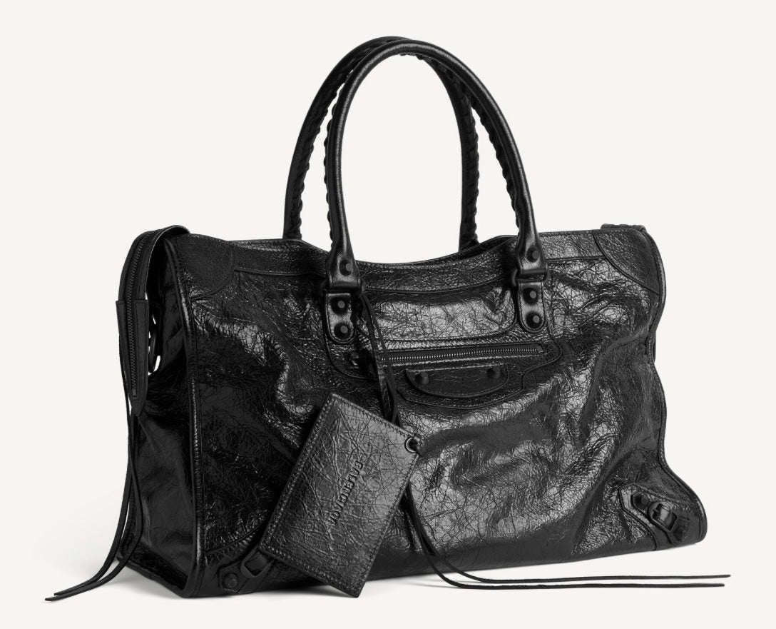 Black Balenciaga City bag in textured lambskin, with rounded handles, a front zip pocket, dark hardware, long leather ties and a hanging rectangular accessory against a pale background.