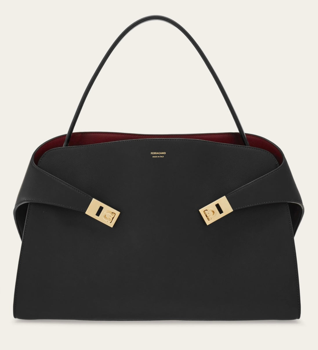 Front view of a black Ferragamo Hug bag with a single arched handle, wraparound straps, gold-tone fittings and a burgundy interior against a pale background.